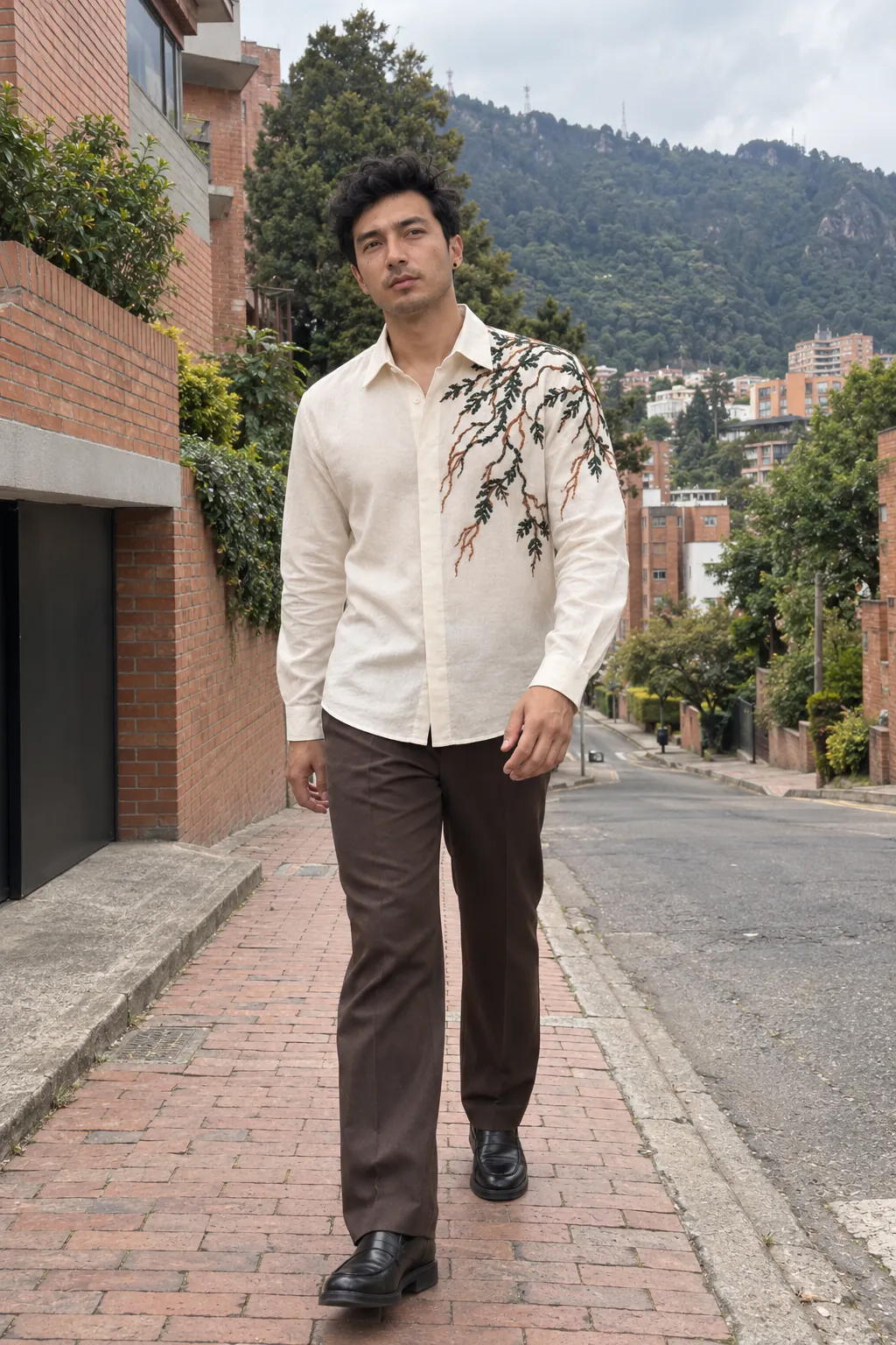 Agui, de cuerpo entero, en una calle de Chapinero Alto, en Bogotá, con una camisa marfil de cuello en punta con un bordado de ramas en verde y cobre en el hombro izquierdo, y pantalón café de sastrería. Imagen de campaña.