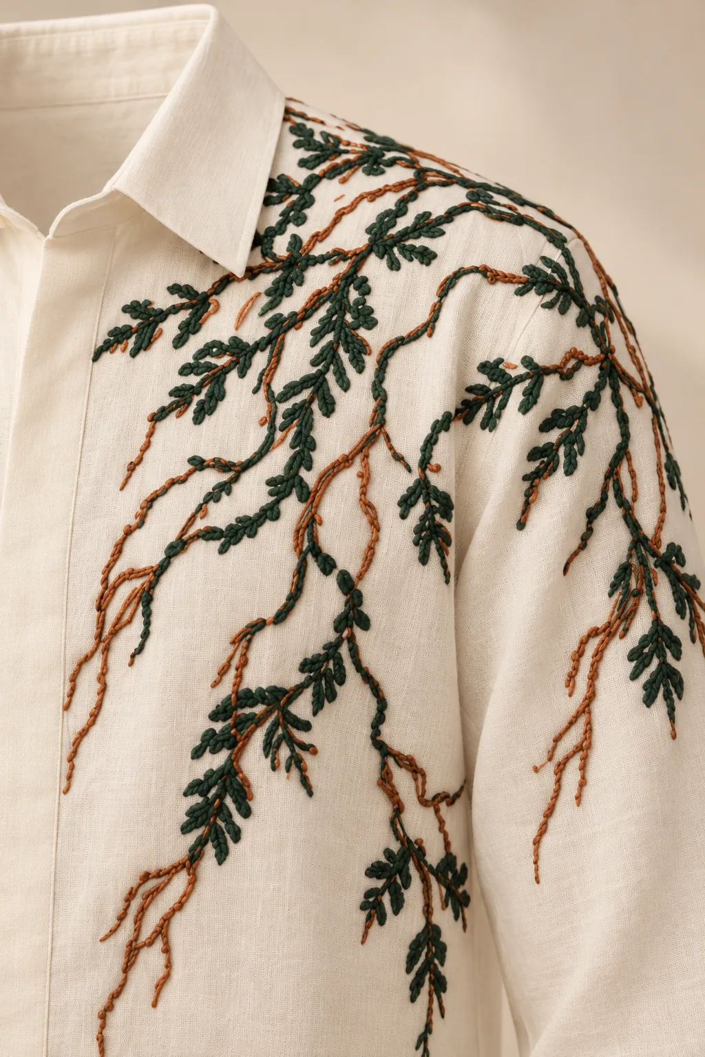 Detalle del bordado de una camisa marfil de cuello en punta con un bordado de ramas en verde y cobre. Solo la prenda, sin modelo.