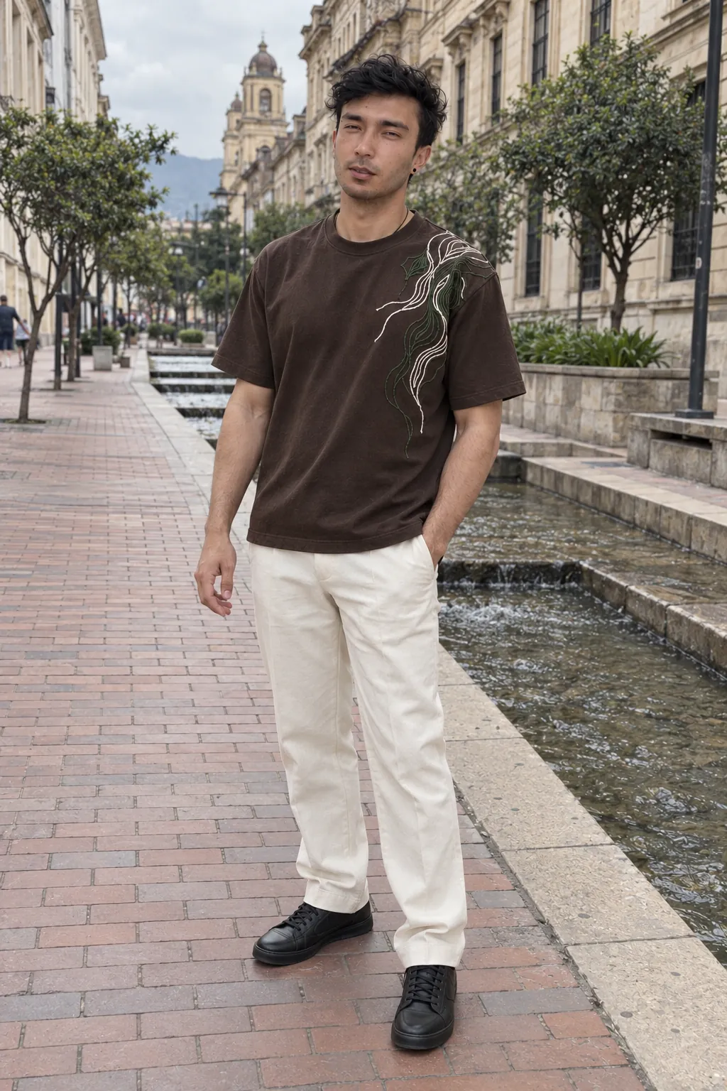Agui, de cuerpo entero, en el Eje Ambiental, en el centro de Bogotá, con una camiseta café de manga corta con un bordado de contornos en marfil y verde en el hombro izquierdo, y pantalón marfil de sastrería. Imagen de campaña.