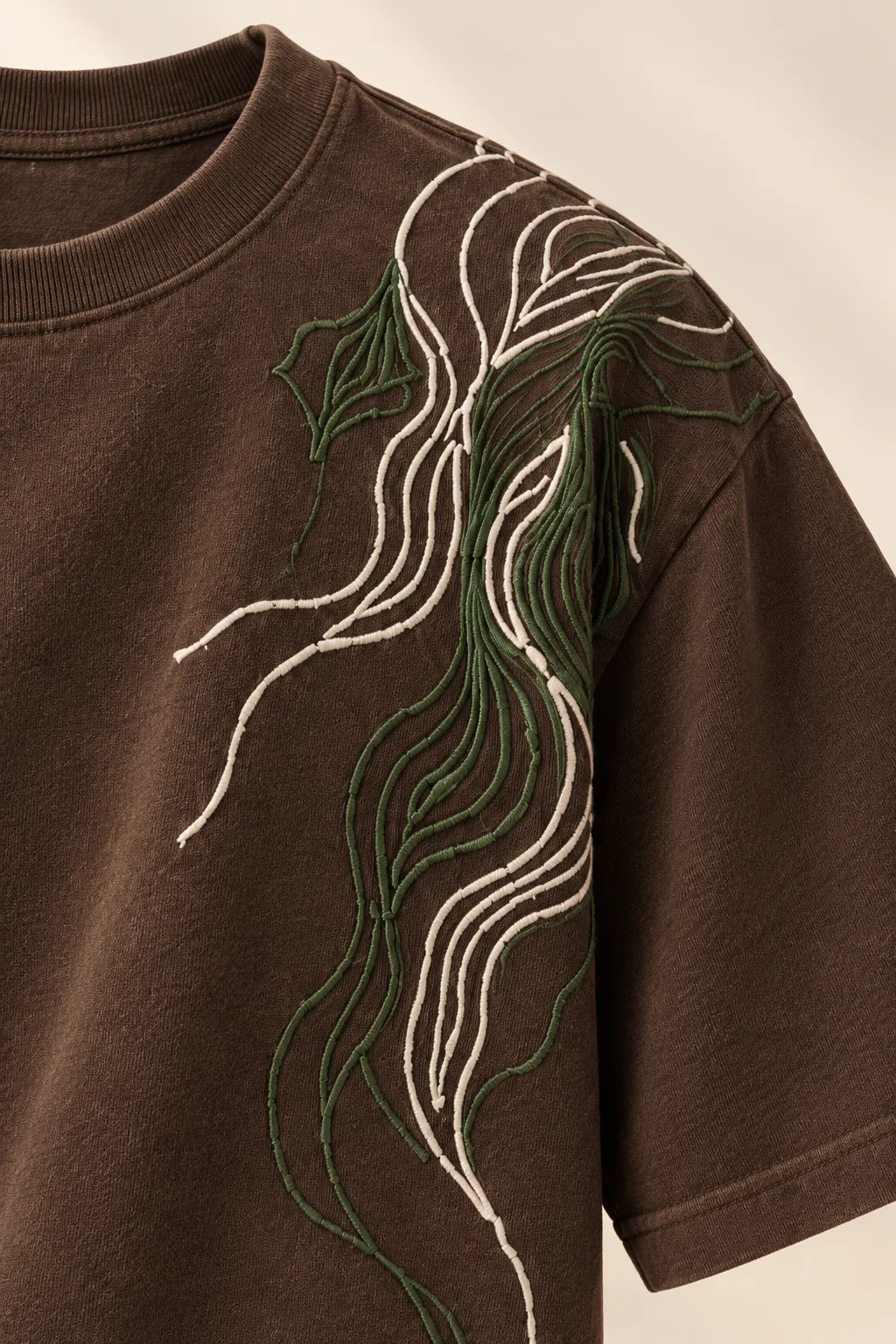 Detalle del bordado de una camiseta café de manga corta con un bordado de contornos en marfil y verde. Solo la prenda, sin modelo.