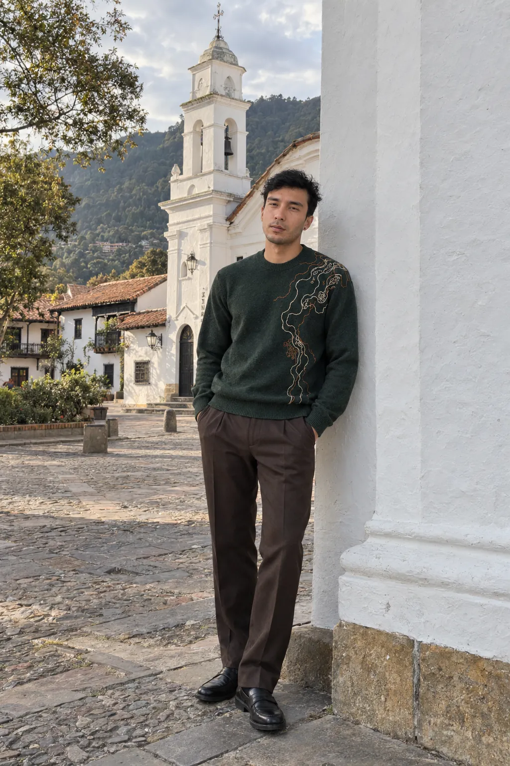 Agui, de cuerpo entero, en la plaza de Usaquén, en Bogotá, con un suéter de punto verde bosque con un bordado de contornos en marfil y cobre en el hombro izquierdo, y pantalón café de sastrería. Imagen de campaña.