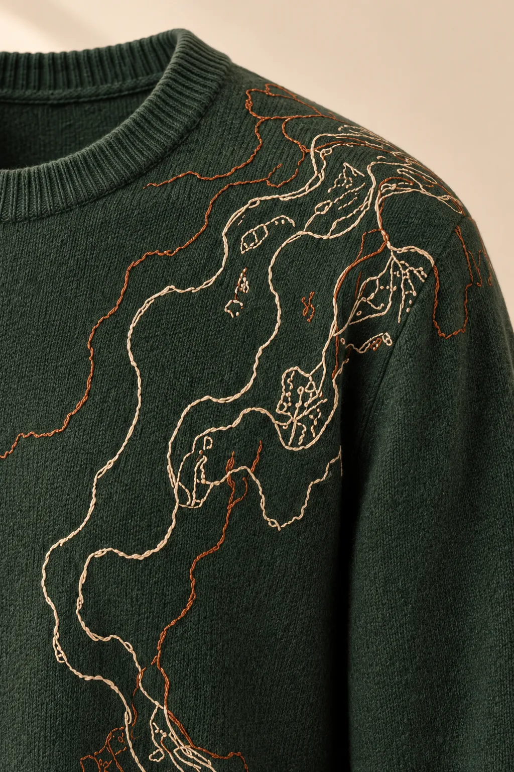 Detalle del bordado de un suéter de punto verde bosque con un bordado de contornos en marfil y cobre. Solo la prenda, sin modelo.