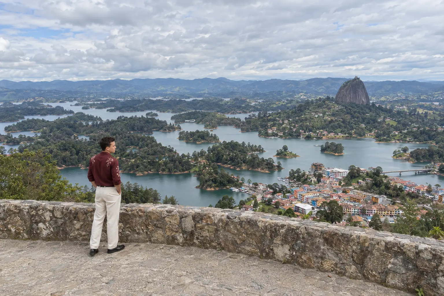 Days, de espaldas a tres cuartos, junto al muro del mirador sobre el embalse de Guatapé, con la camisa vinotinto de flores bordadas. El Peñol al fondo.