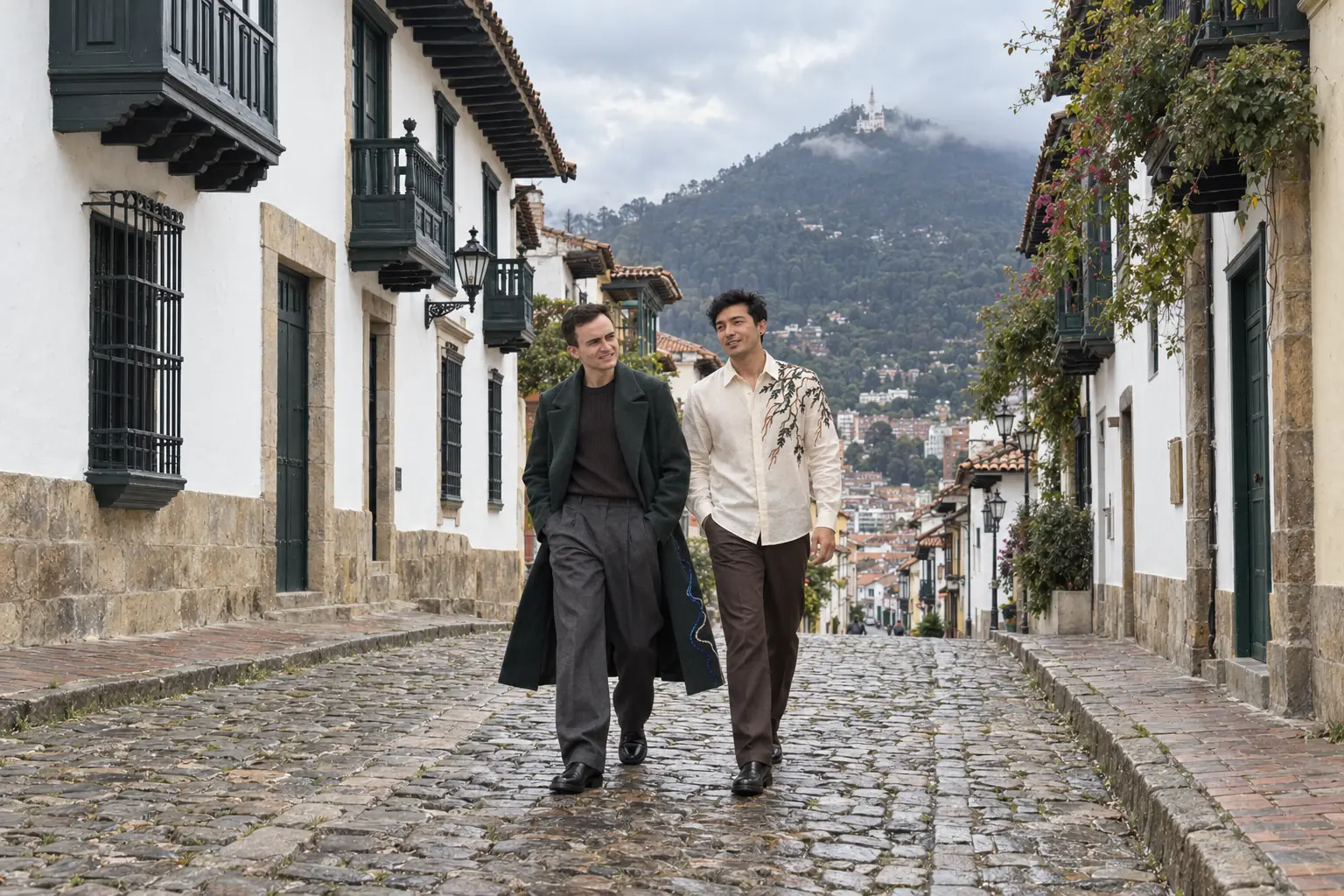 Days y Agui caminan juntos por una calle empedrada de La Candelaria, en Bogotá. Days lleva el abrigo largo verde bosque y Agui la camisa marfil de cuello en punta.